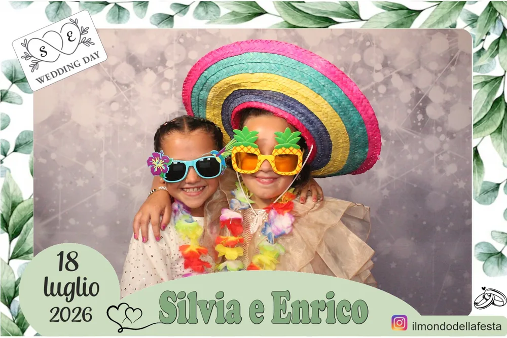 Due bambine con sombrero colorato, occhiali buffi e collane di fiori dentro la cornice a foglie di eucalipto del matrimonio del 18 luglio