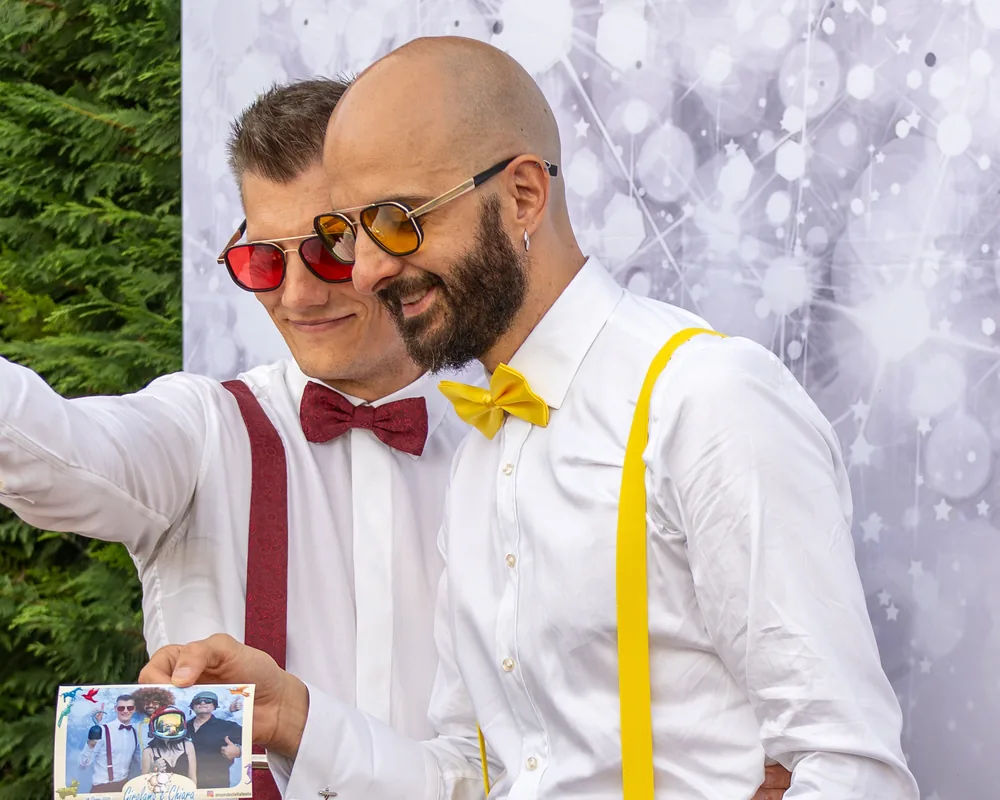 Due invitati in camicia bianca, papillon bordeaux e giallo e occhiali colorati: uno tiene davanti a sé la stampa 10x15 appena ritirata, con la cornice «Girolamo e Chiara, 14 giugno 2026».