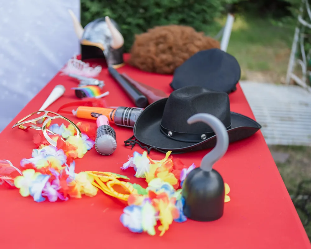 Tavolo con tovaglia rossa coperto di oggetti da indossare: cappello da cowboy, elmo con le corna, parrucca riccia, collane di fiori, occhiali colorati, microfoni finti e un uncino da pirata.